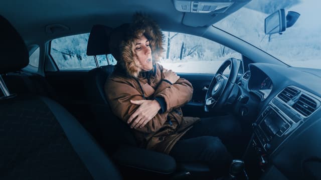 ihminen palelee jäisessä autossa talvivaatteet päällä lumihangessa