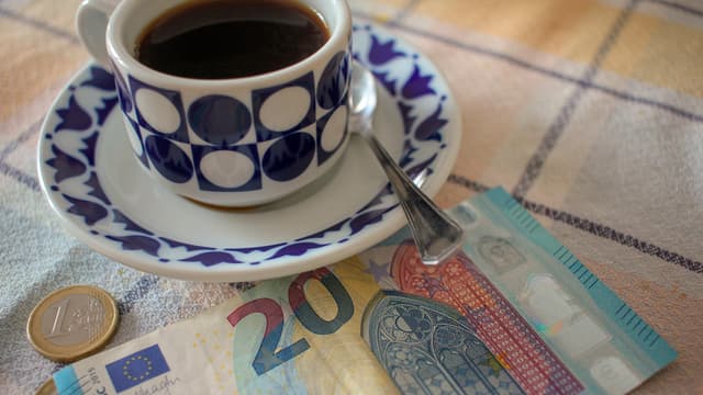 Kahvikuppi, 20 euron seteli ja eurokolikoita pöydällä.